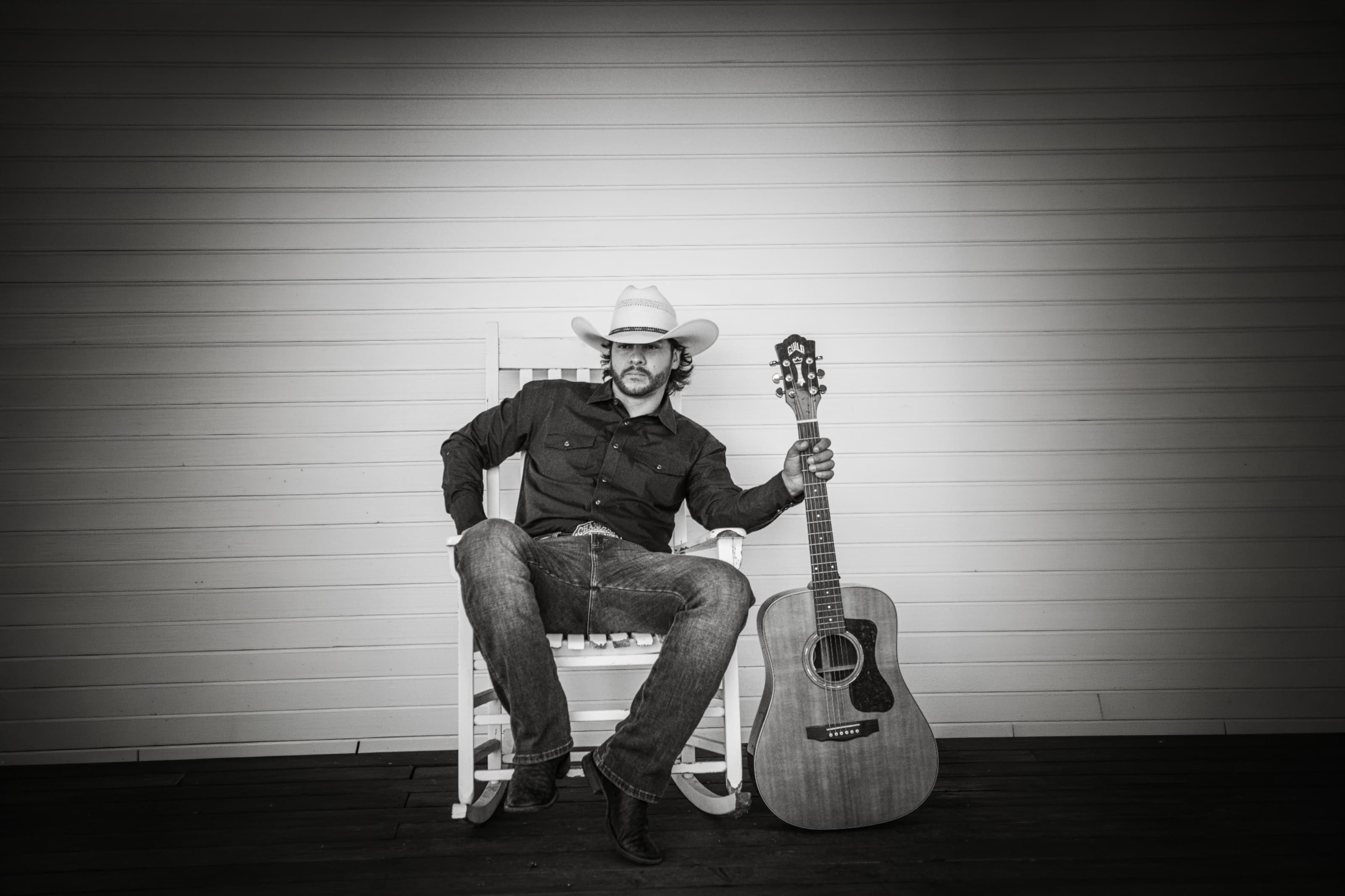 Buck Ford seated in a white rocking chair on a porch, straw hat on, Guild guitar standing beside him